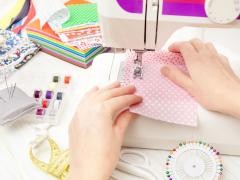 Verschiedene Nähutensilien stehen auf einem Tisch. Darunter auch eine Nhmaschien auf der eine Person gerade einen rosa Stoff näht.