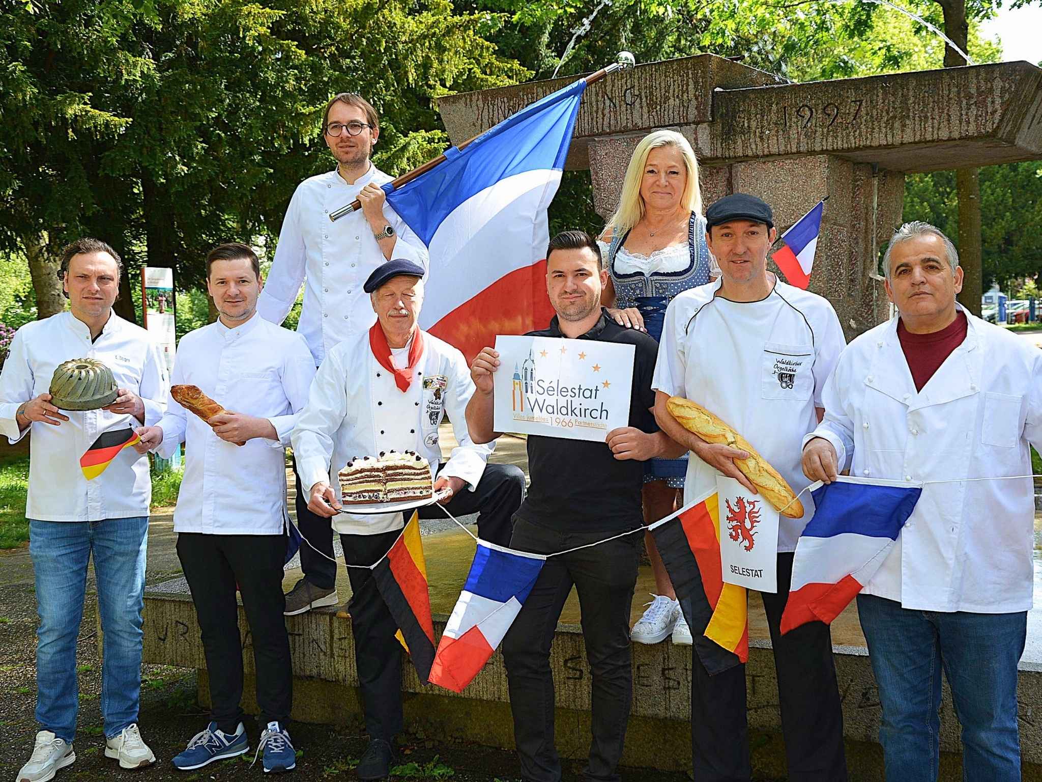 Mehrer Menschen in Kochschürzen, Dirndel oder weißem Hemd posieren für ein Bild. Sie halten Deutschland- und Frankreich-Fahnen.